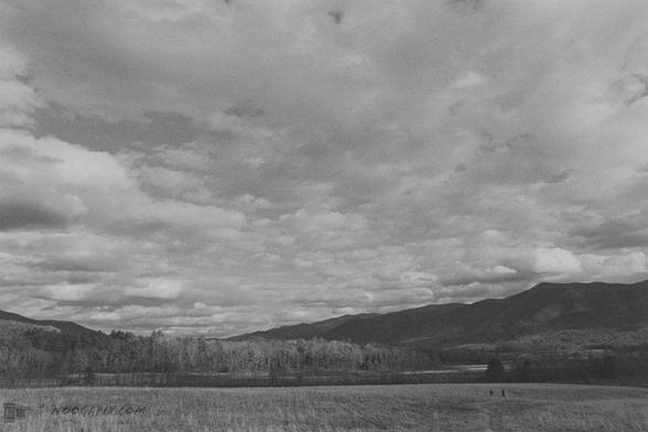 Black and white photo of a field, distant figures, mountains, and a textured cloudy sky