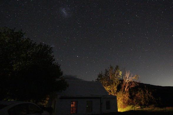 A photo of the night skies in the southern hemisphere 
A small farmhouse with lights on is in the foreground with trees on either side.
A range of mountains is also visible
The tree to  the right is lit by an outside light of the house
A car is barely visible bottom left.
There is a scattering of stars with both Magellanic Clouds visible 
Light pollution is visible on the horizon at right