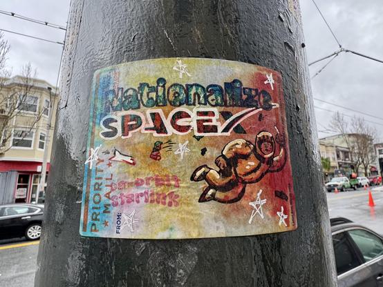 A USPS priority mail label slapped on a green post on Mission Street. It says "Nationalize SpaceX, De-orbit Starlink" and shows Elon Musk in a space suit floating away and flipping off the viewer. It also shows several satellites about to colllide with a space shuttle. The sticker is wet, it is a rainy day, and it has been up for a few months. It's faded, but surprisingly still readable considering it's hand drawn art on an uncoated sticker. 