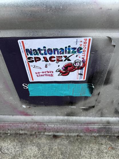 A USPS Priority Mail label with "Nationalize SpaceX" written on it and stuck to one of those grey cans that are filled with dirt and plants and put on sidewalks to prevent folks from being able to use the sidewalk space. This is similar to the other labels in this post, but is a plain white background, instead of the tie-dyed color in behind the art. 