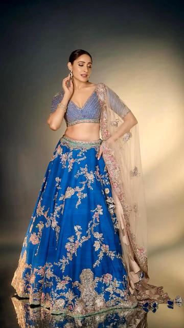 Embodying fairy-tale elegance, Pragya Jaiswal stuns in Dolly J's blue raw silk bridal lehenga, intricately embroidered with gota, zardozi, and thread work. A pale pink tulle dupatta and structured blouse enhance the ensemble's charm. Her bold makeup and royal green jewellery complete the enchanting look.