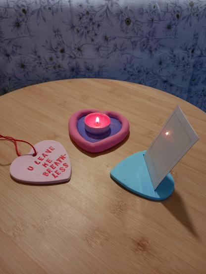 Foto a una mesita de madera donde está un colgante rosa claro en forma de corazón con cuerda de yute roja y el texto "U leave me breathless" ("Me dejas sin aliento", en referencia al episodio de Futurama), un portavelas morado en forma de corazón con los bordes rosas y una vela granate encendida encima y un portafotos de corazón azul con una Polaroid blanca encima en vertical.