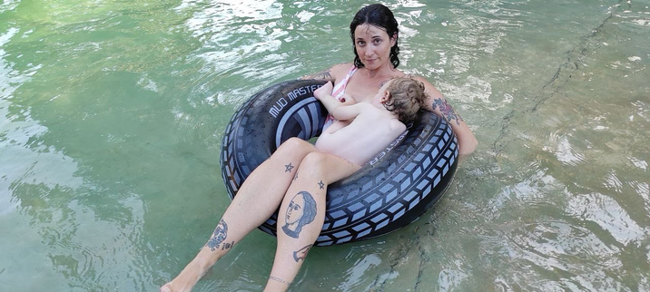 Con un flotador en forma de neumático en una piscina, yo miro a cámara con cara de circunstancias, mi hijo desnudo sobre mi busca la teta apartando el tirante del bañador.