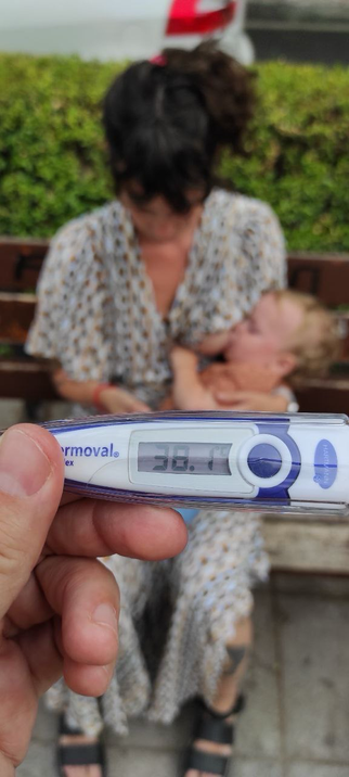 El primer día que mi hijo tuvo fiebre, dar el pecho resultó muy útil. Un termómetro marca 38.1º y al fondo le doy teta sentada en un banco (el fondo está desenfocado).
