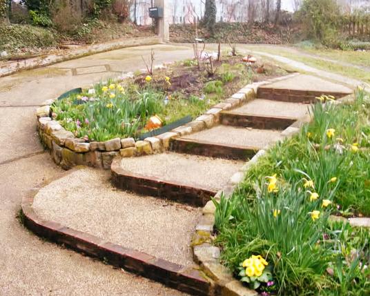 Un escalier extérieur entouré de parterres de fleurs, sur le fond et la gauche il y a une descente de garage PMR qui permet de faire le même trajet.