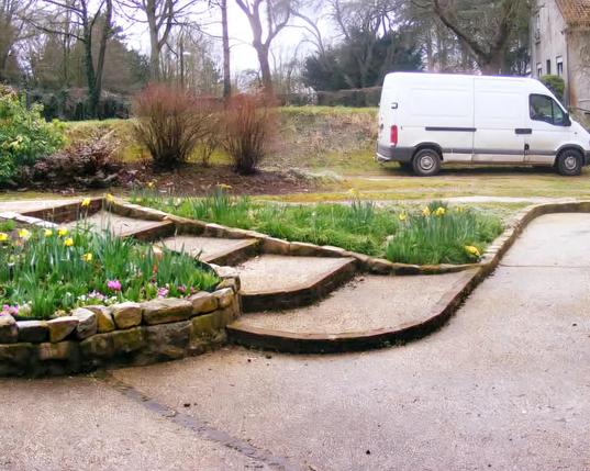 Un escalier extérieur entouré de parterres de fleurs, sur l'avant et la droite il y a une descente de garage PMR qui permet de faire le même trajet. Au fond il y a des arbres et sur la droite un camion et derrière lui une maison.