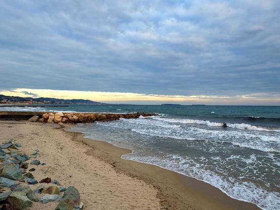 A beach scene featuring long waves lapping against the shore, with a rocky breakwater in the foreground. The sky is overcast, and two islands and distant hills are visible along the coastline.