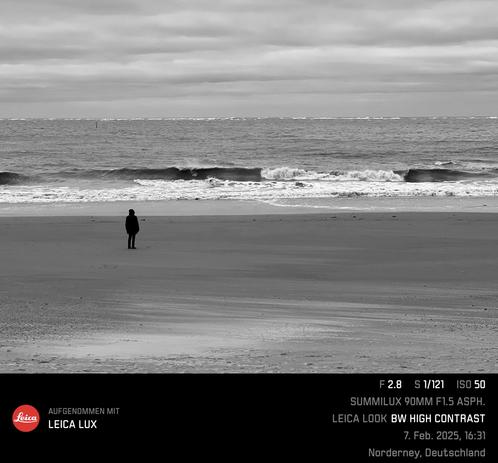 Schwarz-weiß Bild mit hohen Kontrast, ein Mensch an einem ansonsten leeren Strand, den Blick aufs Meer gerichtet, wo eine Welle gerade zu brechen beginnt