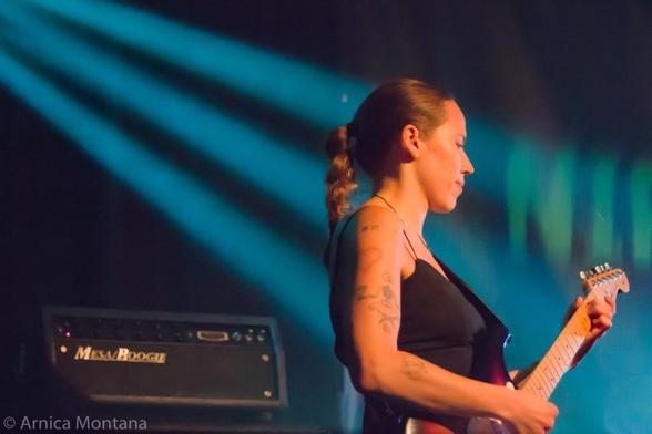 A woman with tattoos plays an electric guitar on stage, illuminated by blue lighting. An amplifier is visible in the background.