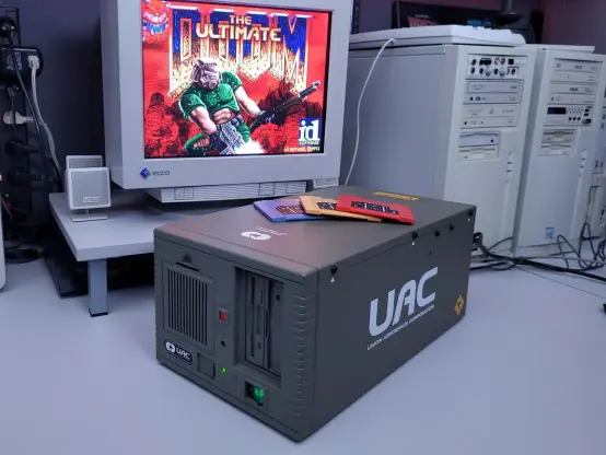 An old PC is shown. 
What is called a 'retro battlestation' because of the age (a 1990s PC) and the utilitarian look to it. It is all white, the monitor is a CRT square shape. The focus on the photo is the 'UAC' computer. This is a modified desktop PC to look like it is from the old DOOM PC games. It is dark gray and has UAC on the site in white. 

Blue, tan and red floppy disks one of each, sit on the top of the computer. 

A static image of THE ULTIMATE DOOM is shown on the monitor. 