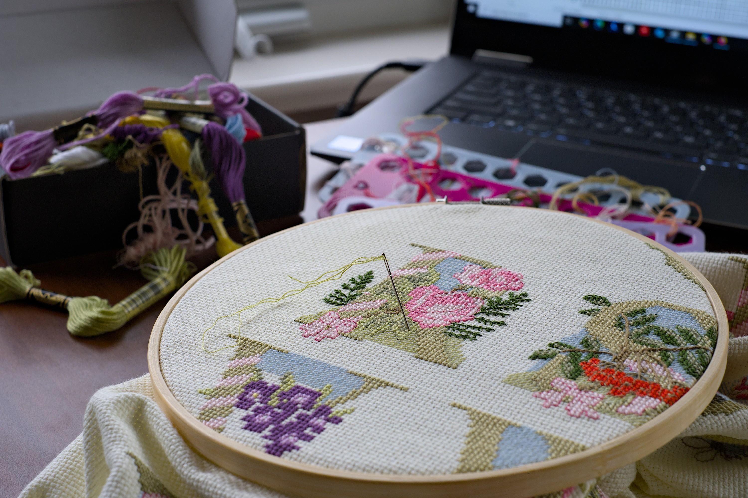 Cross-stitched letter R with a colorful ornament of roses, in a round stitching frame. A needle with a light green thread is stuck into it. The frame is placed on the table in front of a set of colorful threads and an open laptop in the background.