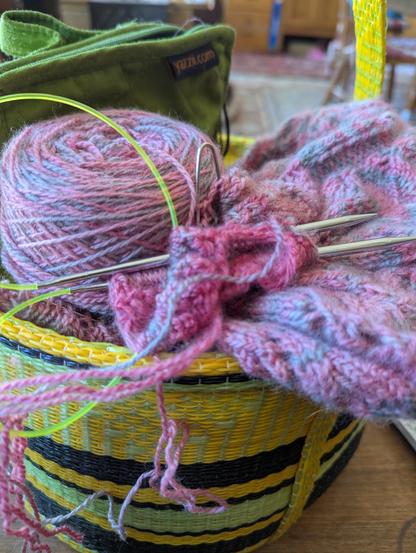 A cake of soft pink and grey yarn sits with lace knitting in the same yarn on circular needles, all in a yellow, black and green striped basket.