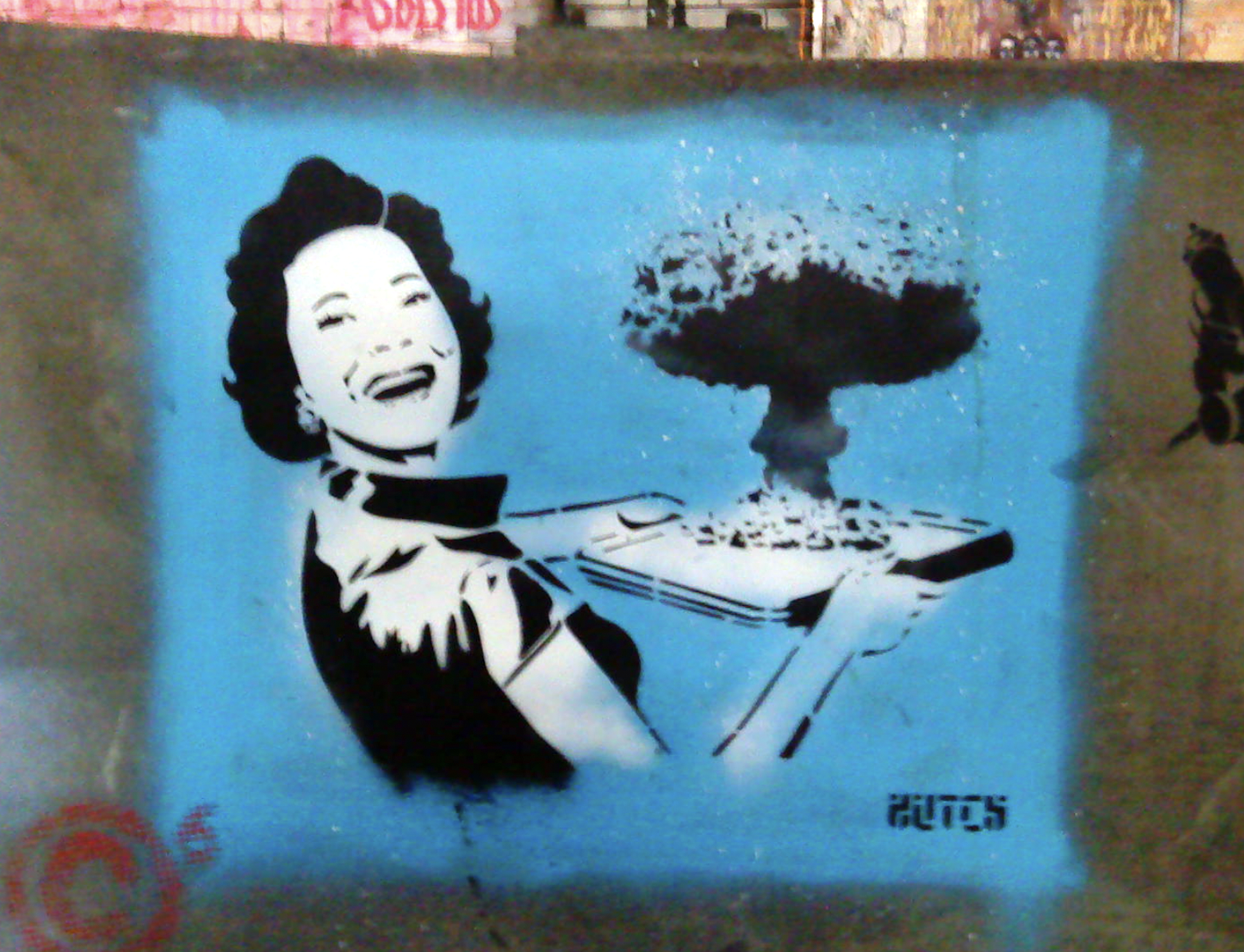 Photo of street art on a concrete wall.  Against a blue background a smiling 1950s housewife in black and white holds a serving tray containing a mushroom cloud.