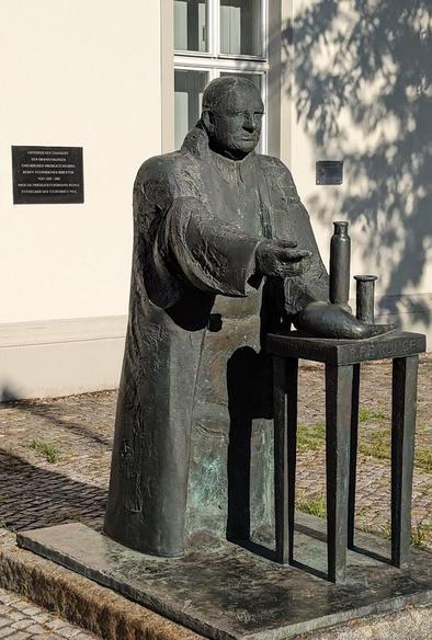 Zu sehen ist eine Bronzestatue eines Mannes. Die Statue steht im Freien vor einem Haus. Der Mann trägt einen Kittel und steht vor einem kleinen Tisch mit Laborzubehör.