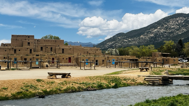A panoramic photo of Taos Pueblo, one of the oldest continuously inhabited pueblos in the country. There is a river in the foreground, a dirt path in the middle ground. In the background to the left is a traditional multi-story adobe structure. To the right, the foot of a mountain seems to converge with the building structure. All of this is set against the backdrop of a blue sky with stratus and cumulus clouds. 