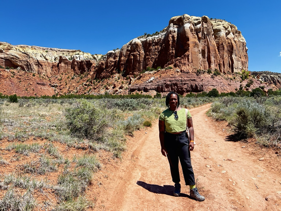 A panoramic photo of Osa Atoe standing on a red dirt path surrounded by desert brush. In the background is an epically large and colorful rock formation against a bright blue cloudless sky. The location is the Ghost Ranch in Abiquiu, NM. 