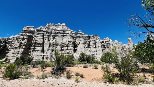 A panoramic view of La Plaza Blanca in Abiquiu, NM. The foreground shows dry soil with scattered desert brush. The middle ground shows an epically large grayish white rock formation. The background is a bright blue cloudless sky. 