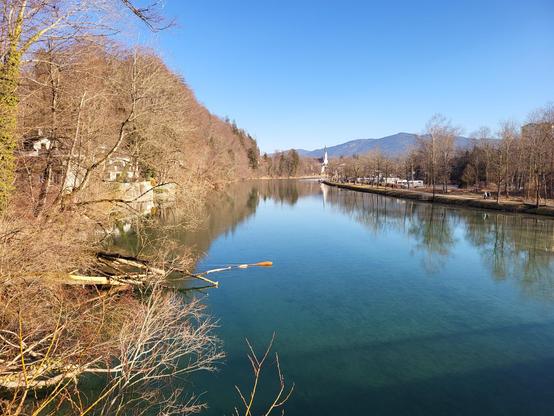 A serene river flows gently through the landscape, flanked by lush trees and a distant building in the background. The scenery is peaceful and picturesque, with the calm waters reflecting the surrounding greenery. The sky above is a beautiful shade of blue, dotted with fluffy white clouds. The setting evokes a sense of tranquility and natural beauty, with the riverbank providing a perfect spot for relaxation and contemplation. The overall scene is a harmonious blend of water, trees, and architecture, creating a peaceful oasis in nature.