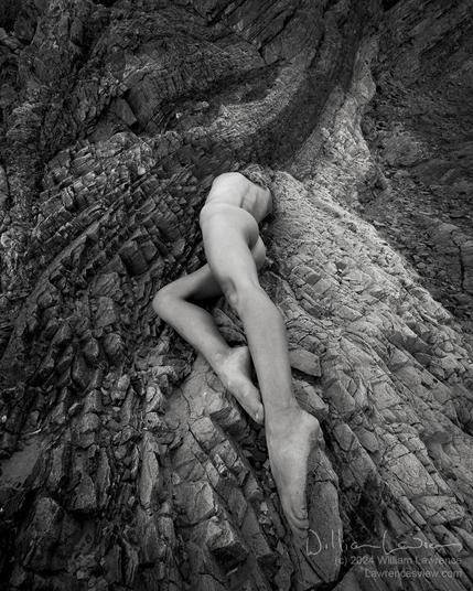 Black and white photograph of model Vivian Cove, posing nude on a rock formation with various toned strata that curves upwards in the distance (it reminded me of a wave).  Vivian lies on the rock facing away from the camera, with her legs to the camera in a very wide angle shot, so her legs are long and the rest of her body foreshortened, with her body making a lovely line leading along the curve of the rock.