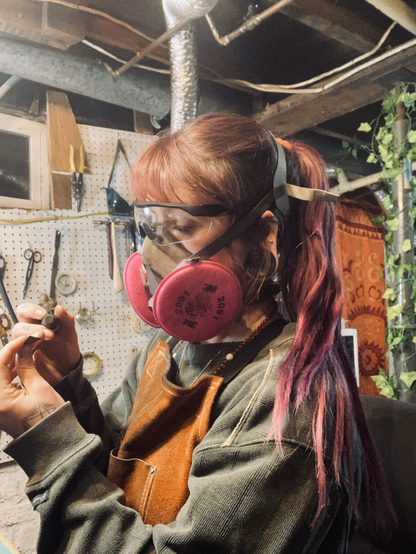 A woman, shown from side profile, holds up a small piece of metal being polished. Her magenta hair is held in a ponytail for safety, and she is also using. A respirator and safety glasses. A dark green sweater and craftsman apron are also being worn. In the background is a white pegboard with an assortment of tools on it. 