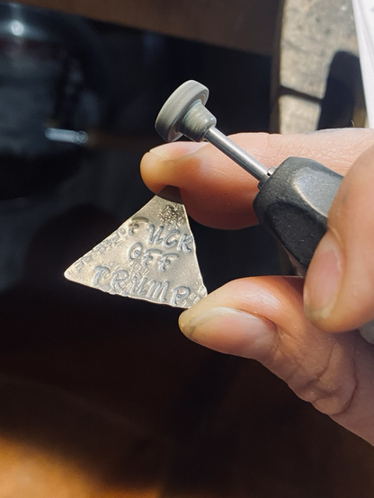 A close up of what the artist was working on, a triangular piece of sterling silver metal that has the phrase “Fuck off Trump” being held with the artists middle finger and thumb while also holding the polishing tool with circular polishing attachment.