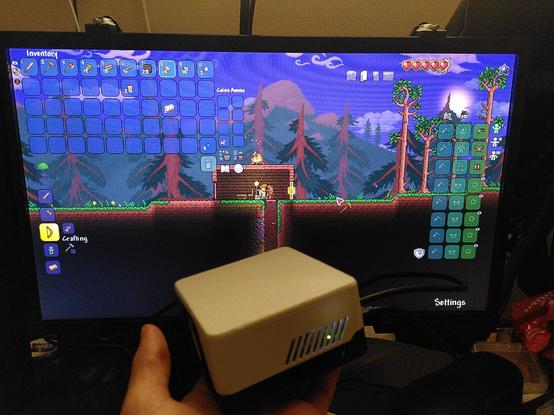 Ruka drží Raspberry Pi 5 v custom krabičce. V pozadí hra Terraria