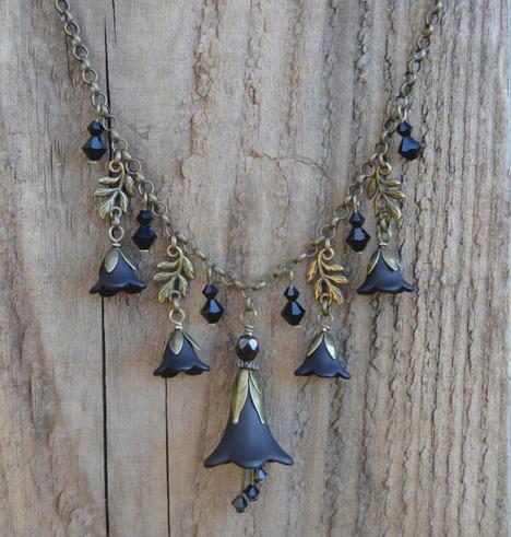 Black flower necklace with a large central flower, and smaller flowers, leaves, and crystal pendants hanging either side from a bronze chain. 3 small crystals dangle underneth the largest flower pendant. Background is faded rough wood.