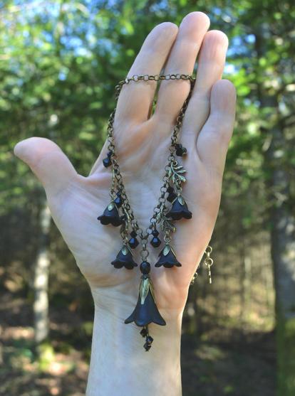 Necklace of dangling black flowers and crystals from a bronze chain, held in the palm of an upright hand. Dark pine trees and appled sunlight behind.