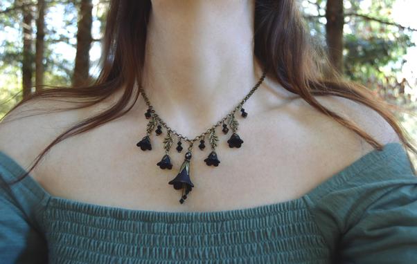 Gothic black flower necklace worn by a figure in a green dress with long reddish hair, standing in a forest. The necklace is V shaped, a large black flower with dangling crystals below, and smaller flowers and crystals hanging from the chain on either side.