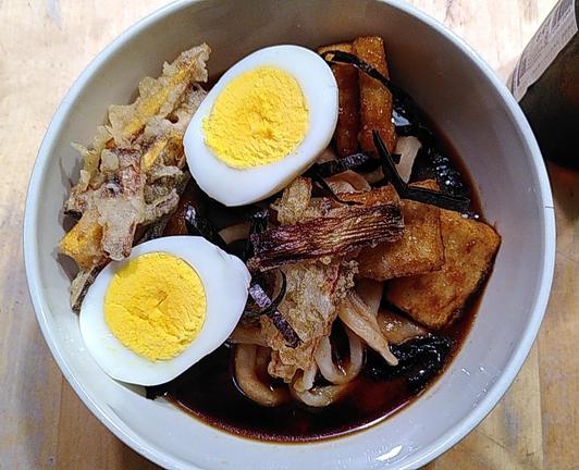 Un bol avec des Udon trempant dans un bouillon, par dessus des rectangles de tofu frits, 2 demis oeufs durs, des tempuras de légumes, et du nori.