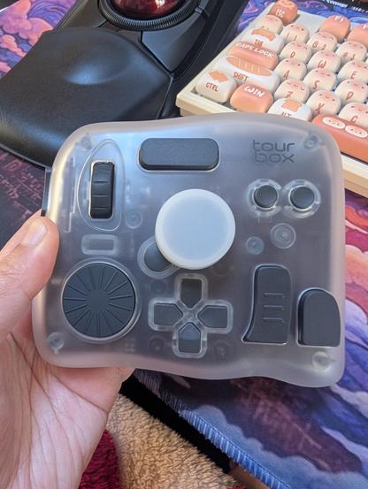 Photo of a left hand holding a Tourbox Elite Plus with a trackball and bun themed keyboard in the background.