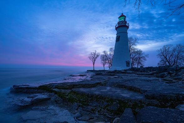 A lighthouse along the shore at sunrise. Pre-sunrise blue tones are prominent with the first warm light just starting to show in the distant clouds.