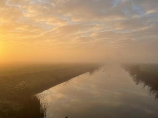 Morning glow over a countryside stream of water with a bank on both sides. The water is reflecting the clouds. The sunrise is colouring the clouds yellow, and it's a little foggy. It's a beautiful crispy morning. 