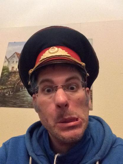 Selfie von mir mit Grimasse und einem Hut der sowjetischen Miliz. Ich trage eine randlose Brille und einen blauen Kapuzenpullover.