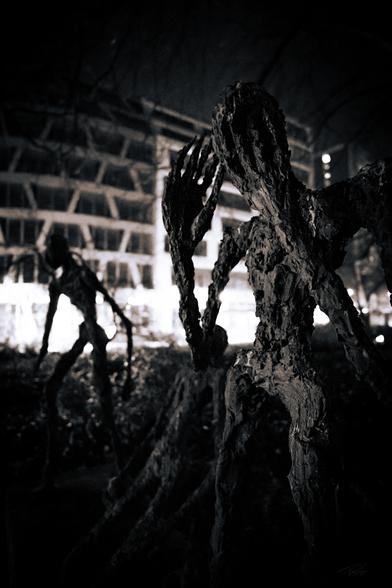 A black-and-white photograph of eerie, textured sculptures at Hackescher Markt in Berlin. The foreground features a dark, humanoid figure with an outstretched hand partially covering its face, while another similar figure stands in the blurred background. Bright city lights contrast with the haunting presence of the statues, creating a dramatic and mysterious atmosphere.