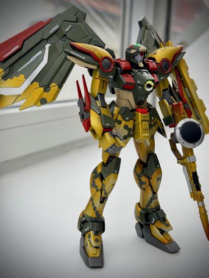 A detailed model of a mech with prominent wings, featuring a camouflage paint scheme in yellow and green. It holds a weapon and has a complex design with red and gray accents. The background is minimal, emphasizing the model's intricate features.
