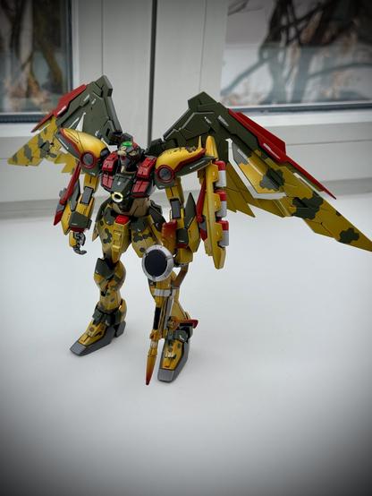 A detailed model of a robot with large, colorful wings. The robot features a camouflage design, predominantly in green and yellow, with red and silver accents. It stands on a white surface with a window in the background.