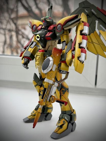 A stylized action figure featuring a humanoid robot with striking yellow, green, and red colors. It has large wings and holds a weapon, set against a softly blurred background with natural lighting.