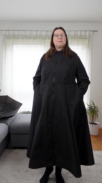 Full body image of a woman in a black raincoat. Hands are in the pockets.