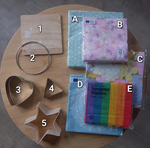 Foto con moldes y paquetes de servilletas con estampados sobre una mesita. A la izquierda de la foto se encuentran diferentes moldes para hacer formas:
1 - Cuadrado (es una tabla de madera, no un molde)
2 - Círculo (aro de metal)
3 - Corazón
4 - Triángulo
5 - Estrella.
A la derecha, las servilletas:
A - Fondo verde pastel con círculos que se solapan estilo años 60
B - Estampado de flores en tonos rosados y malvas pastel como pintadas en acuarelas (el paquete es más pequeño)
C - Estampado de flores y hojas de varios tipos con colores pasteles pero planos (amarillo, rosas, lilas, verde césped)
D - Fondo azul claro con un motivo circular en el centro de la imagen con muchos detalles que podrían recordar a época victoriana
E - Servilletas de un tamaño ligeramente menor rematadas en troquel de línea de semicírculos con franjas de los colores de la bandera LGTBIAQ+ (morado, azul, verde, amarillo, naranja y rojo).