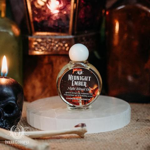 A small circular vial of yellowish Midnight Ember Night Ritual Oil sits on a selenite gemstone disc. It's surrounded by animal bones and a burning black skull candle. It has a spherical white cap and a label that lists its ingredients: "Apricot Kernel Oil, Fragrance Oil" which includes Bergamot, Pine, Warm Spices, and Vanilla Musk. This is an exclusive product from Inked Goddess Creations.
