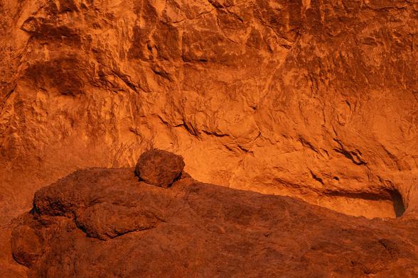 A boulder setting on a outcropping against a cliff wall in the background. The image is warm toned with the cliff wall behind bright and the rock in the foreground darker for contrast.