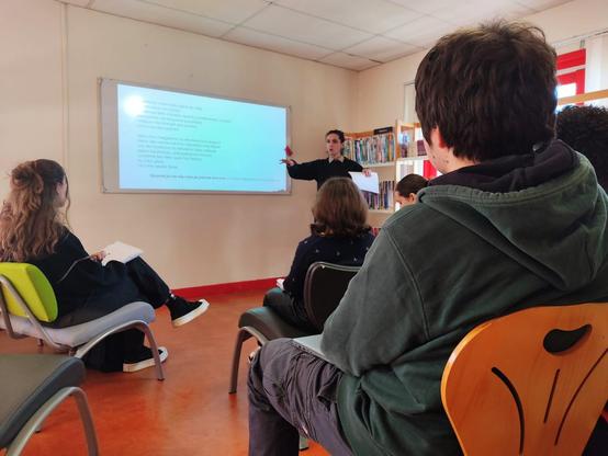 Des lycéens assis dans une salle face à un tableau blanc. Certains prennent des notes. Au bas du tableau, on peut lire le nom de Camille Readman Prud'homme. Debout, Miel Pagès en train de parler au public.