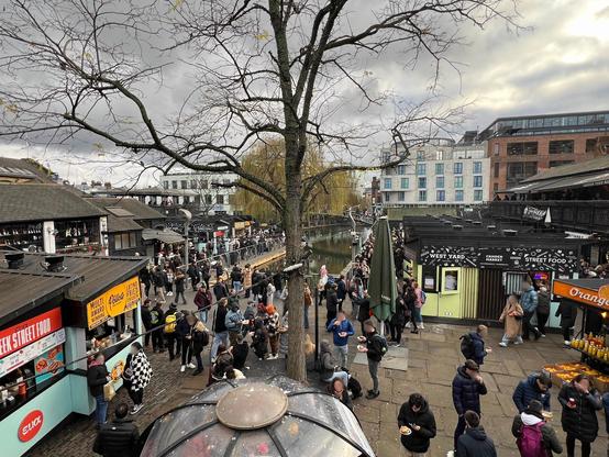 Marché West Yard animé : Une perspective animée sur les stands de street food du West Yard à Camden Market, sous un ciel bleu parsemé de nuages. A bustling view of the West Yard street food stalls in Camden Market, under a partly cloudy blue sky.