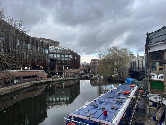 Vue sur le canal Camden Lock : Une vue tranquille sur le canal entouré de bâtiments modernes et d'arbres, avec un bateau amarré sur l'eau calme. A serene view of the canal flanked by modern buildings and trees, with a boat docked on the still water.