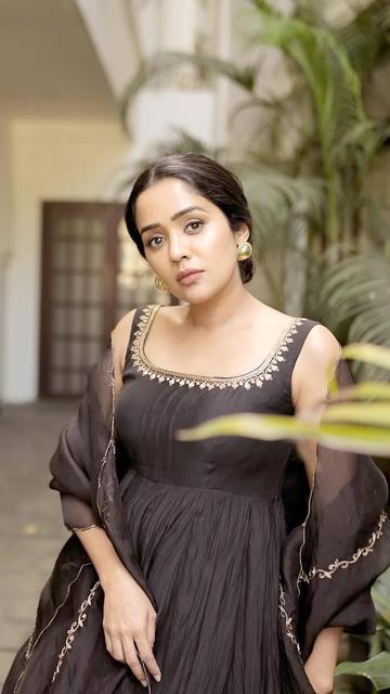 Ananya's black monochrome ensemble is a stunning blend of traditional and modern elements. The sleeveless top features a wide neckline, intricate golden embroidery, and sequins, flaring gracefully below the knee to create a flowing silhouette. She pairs this with matching black flared pants and a sheer dupatta with scalloped golden edges. Statement golden accessories, a sleek bun hairstyle, and elegant peep-toe sandals add the perfect finishing touches to her sophisticated look.