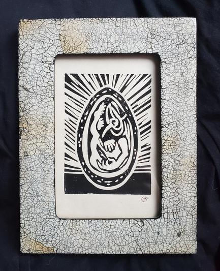 Une lino encadrée. Le cadre est blanc avec des craquelures noires comme de la coquille d'oeuf. La lino représente un oeuf en coupe, dans lequel on voit Jean-Pigeon, un personnage humanoïde tout nu portant un masque d'oiseau, installé jambes en l'air et contemplant une plume qu'il tient dans sa main.