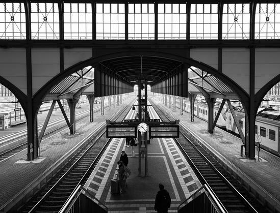 Motiv:
Das Bild zeigt einen Bahnhof mit symmetrischer Architektur. Eine große Stahl- und Glasstruktur überspannt die Bahnsteige. Züge stehen an den Gleisen. Eine Uhr und digitale Anzeigetafeln befinden sich in der Mitte. Wenige Personen bewegen sich auf dem Bahnsteig, einige mit Gepäck. Der Blick führt entlang der Gleise in die Ferne.

Stimmung & Wirkung:
Die Komposition betont die Tiefe des Raums. Die leeren Gleise und die reduzierte Anzahl an Personen lenken den Fokus auf die Architektur. Die Perspektive erzeugt eine Richtung ins Bildzentrum.

Technische Details:
Bildformat: Architekturfotografie mit dokumentarischem Charakter.

Farbe: Schwarz-Weiß.

Schärfentiefe: Hohe Tiefenschärfe, alle Elemente sind klar erkennbar.

Lichtverhältnisse: Gleichmäßige Ausleuchtung durch Tageslicht, gestreut durch Glasflächen.