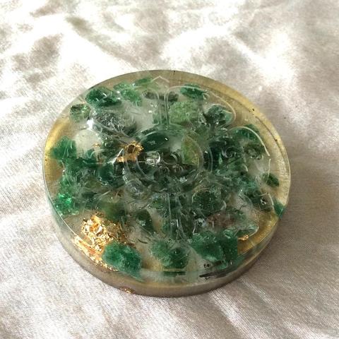 Een kleine orgonite oplaadschijf van 5 cm doorsnee, met groene aventurijn edelstenen en bladgoud. De schijf bevat het Tibetaanse mantra-symbool "Om Mani Padme Hum" en is handgemaakt in Haarlem. Het product absorbeert elektromagnetische straling, bevordert positieve energie en helpt bij fysieke en emotionele balans. Geschikt voor het opladen van edelstenen, voedsel, dranken en medicijnen.
