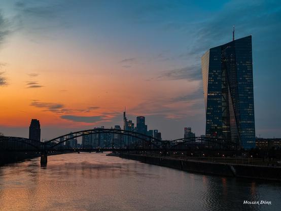 Eine malerische Abendaufnahme von Frankfurt am Main, die die imposante Skyline der Stadt und den markanten EZB-Turm in sanftes Licht taucht. Der orangefarbene Himmel spiegelt sich im ruhigen Wasser des Mains, während die Brücke elegant über den Fluss führt und die Lichter der Stadt in der Dämmerung leuchten.